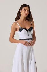 A woman wearing a white sleeveless jumpsuit with thin spaghetti straps. The top has black lace trim along the neckline and a small black bow at the center. The waistline features a black lace band. The jumpsuit has wide-leg pants with pleats and a side slit. The woman has long brown hair, gold earrings, and gold rings on her fingers.